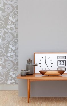 a clock sitting on top of a wooden table in front of a wall with flowers