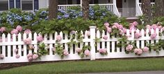 a white picket fence with pink and blue flowers growing on the top, in front of a house