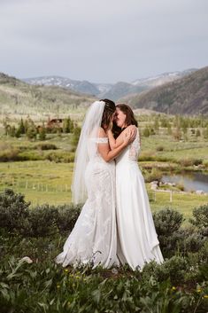 two brides embracing each other in the middle of a field with mountains in the background