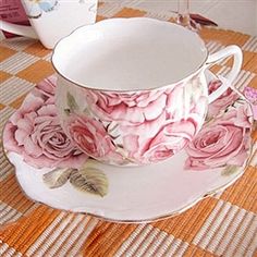 a pink rose tea cup and saucer on a checkered tablecloth with two coffee mugs in the background