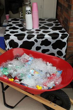 a red wheelbarrow filled with plastic bottles and water in front of a table