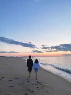 a man and woman walking along the beach with footprints in the sand as the sun sets