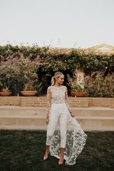 a woman standing in the grass wearing white pants and a sheer top with flowers on it