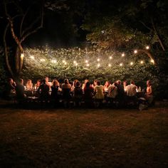 a group of people sitting around a table at night with lights strung from the trees