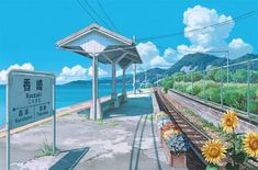 a train station next to the ocean with sunflowers on the platform and a sign