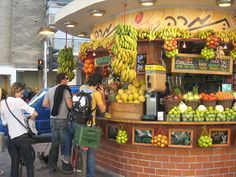 a fruit stand with lots of fruits on display and people standing around the corner looking at it