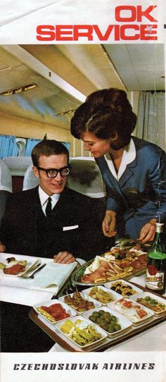 a man and woman sitting at a table in an airplane with plates of food on it