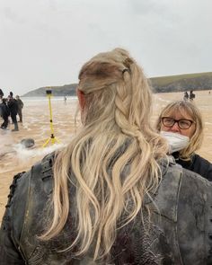 a woman with long blonde hair wearing a leather jacket on the beach while people watch