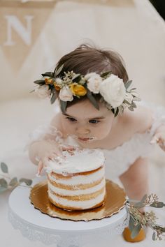 a baby girl wearing a flower crown is looking at a cake