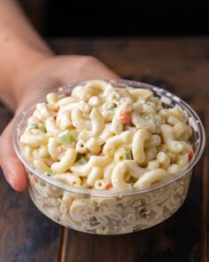 a person holding a plastic bowl filled with macaroni and cheese salad on top of a wooden table