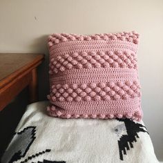 a pink crocheted pillow sitting on top of a white blanket next to a wooden table