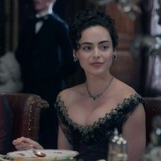 a woman sitting at a dinner table in a black dress with pearls on her neck