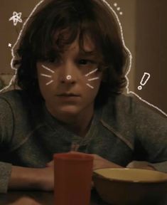 a young boy sitting at a table in front of a cup with cat's eyes drawn on it