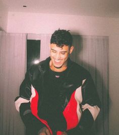 a young man in a black and red jacket smiles at the camera while holding his cell phone