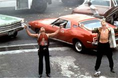 three men standing in front of a red car on the street with their arms up