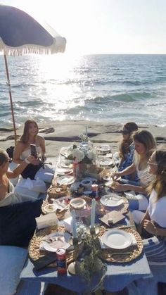 a group of women sitting around a table with plates and drinks on it near the ocean