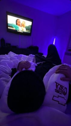 two people laying in bed watching tv with purple lighting on the wall and one person holding a stuffed animal