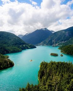 a large body of water surrounded by trees and mountains in the distance with clouds overhead