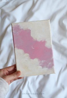 a hand holding a pink and white painting on top of a bed covered in sheets