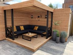 an outdoor seating area with wooden slats and black couches on the floor, surrounded by potted plants