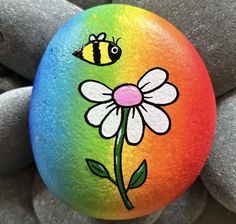 a painted rock with a flower and a bee on it, sitting next to some rocks