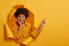 a woman in yellow shirt pointing to the side with her finger and looking through torn paper