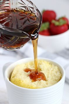 someone is pouring syrup into a bowl of cake with strawberries in the back ground