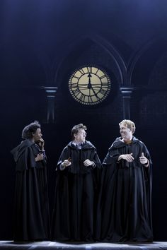 three men dressed in robes stand on stage with a clock tower in the back ground