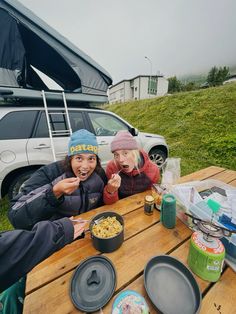 two people sitting at a picnic table with food in front of an suv and camping gear