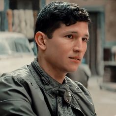 a man with black hair wearing a leather jacket and looking off into the distance while standing on a city street