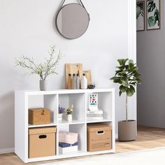 a white shelf filled with boxes and baskets next to a plant on top of a hard wood floor