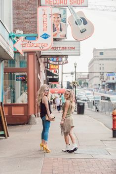 two women walking down the sidewalk in front of a record shop with guitars hanging above them