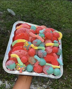 a tray filled with lots of colorful candy on top of a grass covered field next to a person's hand