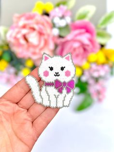 a hand holding a small white cat with a pink bow tie and flowers in the background