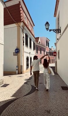 two people walking down a street next to tall white buildings on either side of each other