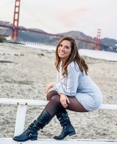 a woman sitting on top of a white fence next to the ocean with a bridge in the background