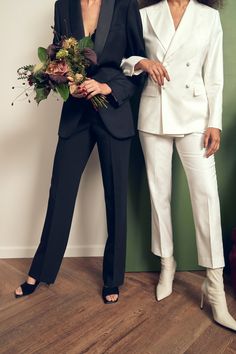 two women standing next to each other in white suits and black shoes, one holding a bouquet