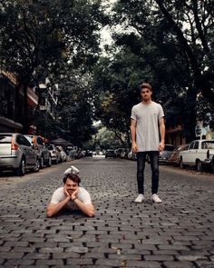 two young men are standing on the street and one is laying on the ground with his head in his hands