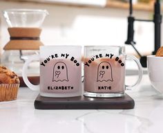 two coffee mugs sitting on top of a counter next to some muffins