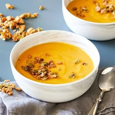 two white bowls filled with carrot soup on top of a blue table cloth next to silver spoons