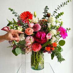 a vase filled with lots of colorful flowers on top of a glass table next to a person's hand