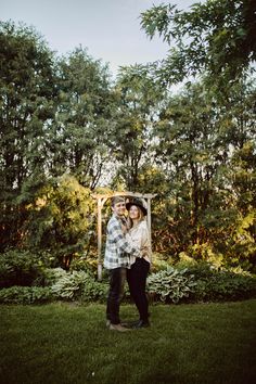 a man and woman standing in the grass with trees behind them, holding an umbrella