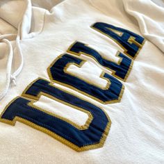 Completely Unworn And New-Without-Tags! Authentic Ucla Collegiate Apparel Purchased From The University Bookstore By Russell Athletic. Thick Mustard Yellow & Navy Embroidered Letters And Off-White/Ivory Heavyweight Cotton/Polyester Fabric Blend. Fleece Lining With Front Pocket - Soft And Cozy! No Damage, Stains Or Signs Of Wear! Measures 20” Armpit To Armpit, 20” Sleeve Length, 25” Shoulder To Bottom Hem. Comes From A Smoke/Pet-Free Home. Varsity Cotton Hoodie With Letter Embroidery, Blue Hoodie With Embroidered Logo For College, Blue College Hoodie With Embroidered Logo, Cotton Varsity Hoodie With Letter Embroidery, Blue Embroidered Logo Hoodie For College, White Sporty Hoodie With Letter Embroidery, White Sporty Hoodie With Embroidered Text, Sporty White Hoodie With Letter Embroidery, White Cotton Sweatshirt With Letter Patch