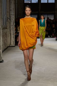 a model walks down the runway in an orange dress with draped over her shoulders, while others look on