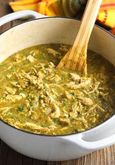 a wooden spoon in a white pot filled with green soup and chicken meat, on a wood table