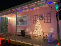 a christmas tree is lit up in front of a pink house with white rocking chairs