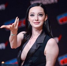 a woman in a black dress holding her hand up to the side while standing at a podium