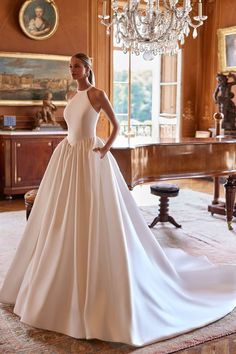 a woman in a white wedding dress standing by a chandelier with a grand piano behind her