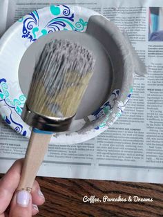 a hand holding a paint brush on top of a paper plate in front of a newspaper