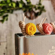 three crocheted snails sitting on top of a book with flowers in the background